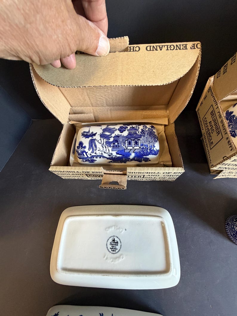 Peut inclure: Beurrier en c&eacute;ramique bleu et blanc avec un motif de paysage, pr&eacute;sent&eacute; dans une bo&icirc;te en carton. Le plat rectangulaire a un couvercle, et la bo&icirc;te est &eacute;tiquet&eacute;e "Churchill of England". Un deuxi&egrave;me plat est visible.
