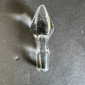 May include: A clear glass bottle stopper with a pointed top. The stopper is shaped like a teardrop and has a smooth, polished finish.