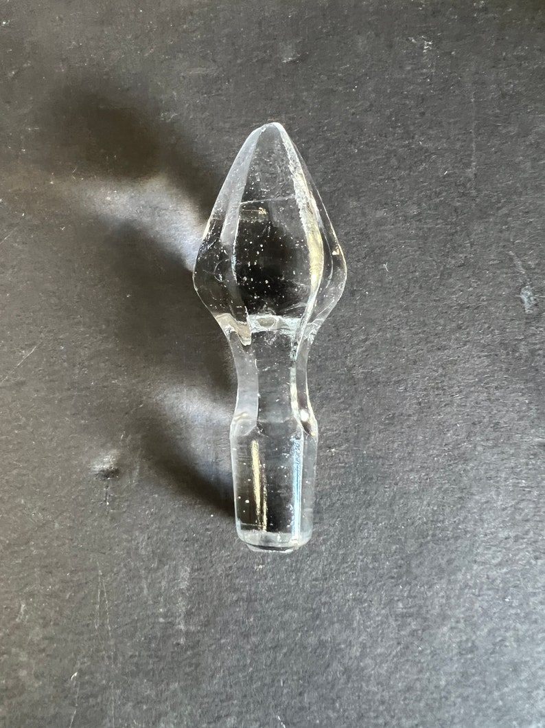 May include: A clear glass bottle stopper with a pointed top. The stopper is shaped like a teardrop and has a smooth, polished finish.