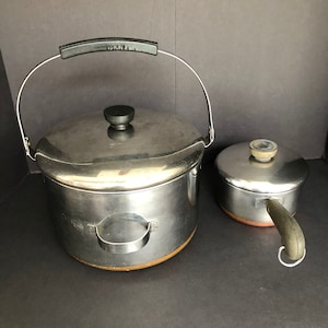 May include: Two stainless steel pots with copper bottoms. The larger pot has a lid and a black handle. The smaller pot has a lid and a black handle with a loop on the end.