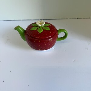 May include: A small ceramic teapot shaped like a red strawberry with a green handle and spout. The lid features a sculpted white strawberry blossom with green leaves centered on top. The body of the teapot has a textured surface with small yellow dots.
