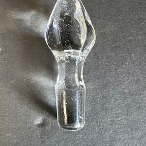 May include: A clear glass bottle stopper with a pointed top. The stopper is designed to fit into the neck of a bottle.