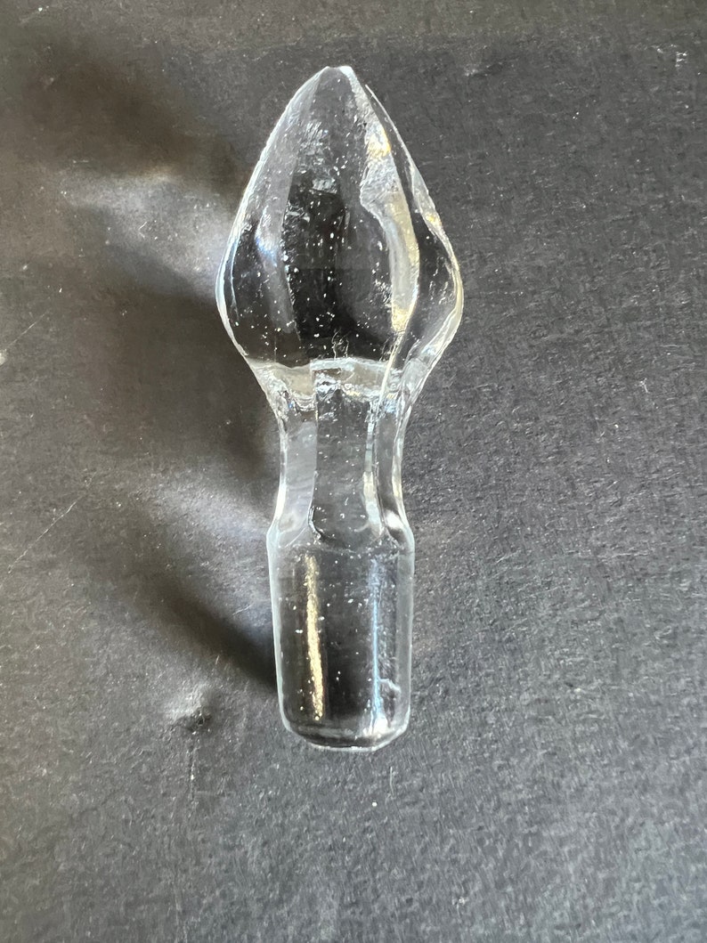 May include: A clear glass bottle stopper with a pointed top. The stopper is designed to fit into the neck of a bottle.