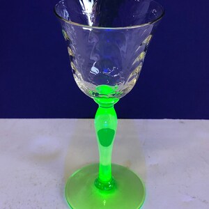 May include: A clear glass goblet with a textured bowl and a neon green stem and base. The goblet has a delicate, ornate design around the rim. The stem and base glow brightly, creating a striking contrast.