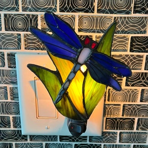 May include: A stained glass dragonfly nightlight plugged into an outlet. The dragonfly is blue and purple with yellow and green accents. The nightlight is lit and casting a warm glow.