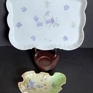 May include: A white porcelain tray with a scalloped edge and floral design. The tray is on a wooden stand. A smaller, leaf-shaped porcelain dish with a floral design is also shown.