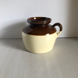 May include: A brown and cream glazed ceramic pitcher with a single handle. The pitcher has a wide mouth and a rounded body.