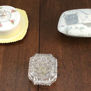 May include: Three decorative boxes, one with a floral design and a yellow lid, one with a white lid and a gray and white design of a building, and one clear glass box with a geometric pattern.