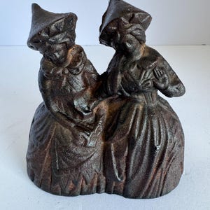 May include: A cast iron sculpture depicting two figures in traditional attire, possibly a historical or folk art piece. The figures are dressed in long skirts and hats, with intricate details in the clothing. The sculpture has a dark brown, aged finish.