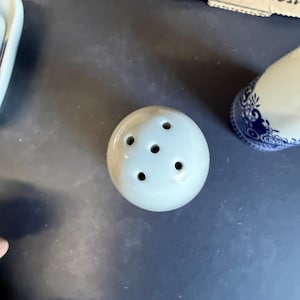 Peut inclure: Un petit sali&egrave;re rond en c&eacute;ramique blanc cass&eacute; avec cinq trous sur le dessus. Le sali&egrave;re fait partie d'un ensemble, avec un &eacute;l&eacute;ment &agrave; motifs bleu et blanc visible en arri&egrave;re-plan. Les articles sont sur une surface sombre.