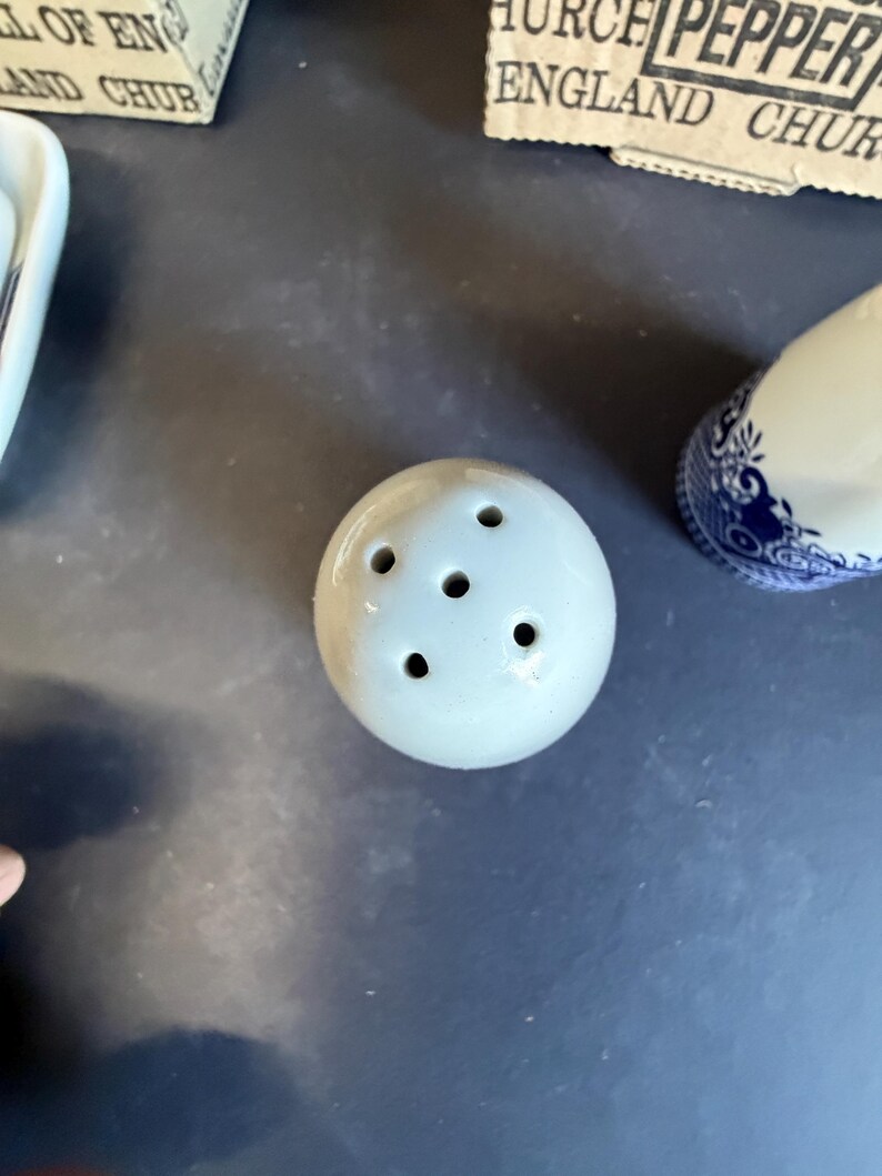 Peut inclure: Un petit sali&egrave;re rond en c&eacute;ramique blanc cass&eacute; avec cinq trous sur le dessus. Le sali&egrave;re fait partie d'un ensemble, avec un &eacute;l&eacute;ment &agrave; motifs bleu et blanc visible en arri&egrave;re-plan. Les articles sont sur une surface sombre.