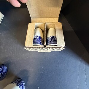 Peut inclure: Deux sali&egrave;res et poivri&egrave;res en c&eacute;ramique blanche et bleue dans une bo&icirc;te en carton. Les sali&egrave;res et poivri&egrave;res ont un motif floral bleu autour de la base et du haut. La bo&icirc;te est ouverte, r&eacute;v&eacute;lant les sali&egrave;res et poivri&egrave;res &agrave; l'int&eacute;rieur.