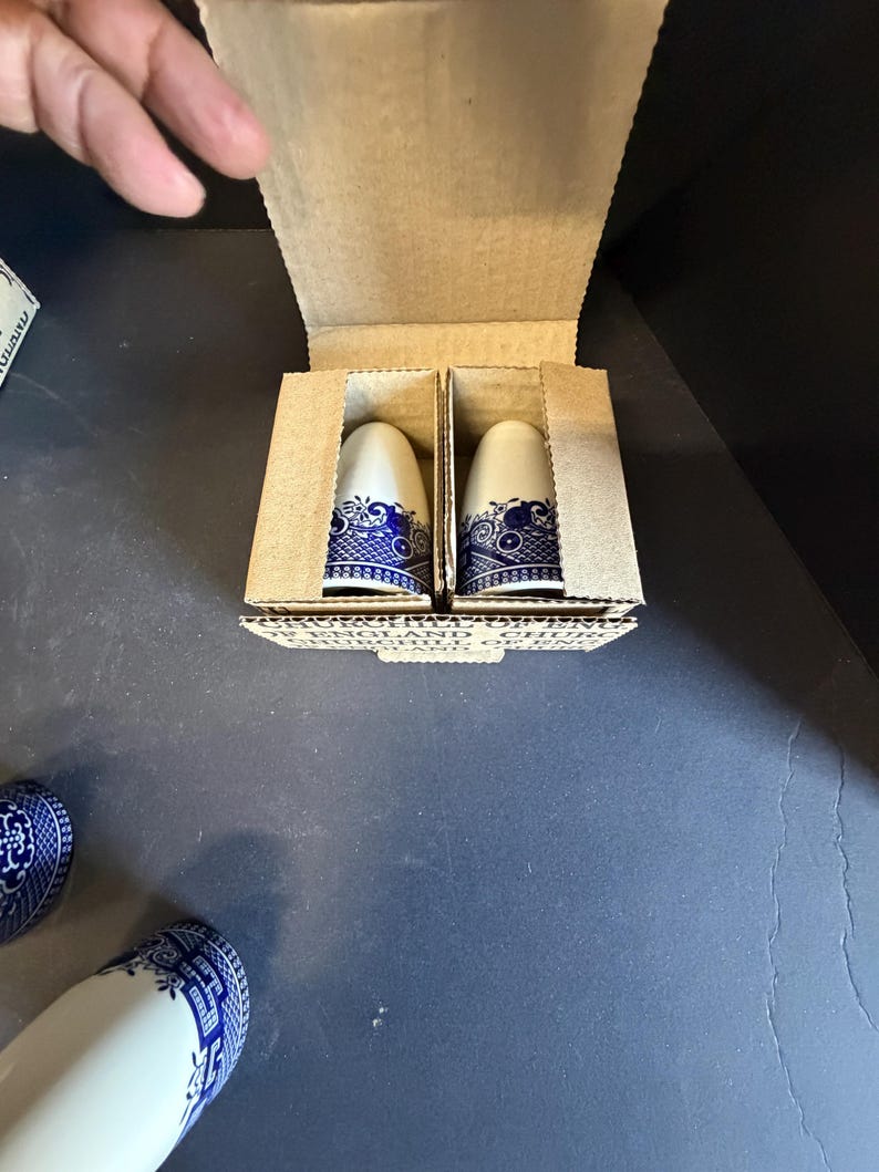 Peut inclure: Deux sali&egrave;res et poivri&egrave;res en c&eacute;ramique blanche et bleue dans une bo&icirc;te en carton. Les sali&egrave;res et poivri&egrave;res ont un motif floral bleu autour de la base et du haut. La bo&icirc;te est ouverte, r&eacute;v&eacute;lant les sali&egrave;res et poivri&egrave;res &agrave; l'int&eacute;rieur.