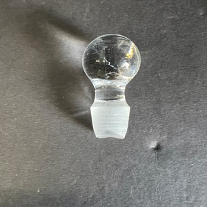 May include: A clear glass bottle stopper with a round top and a flat, rectangular base.