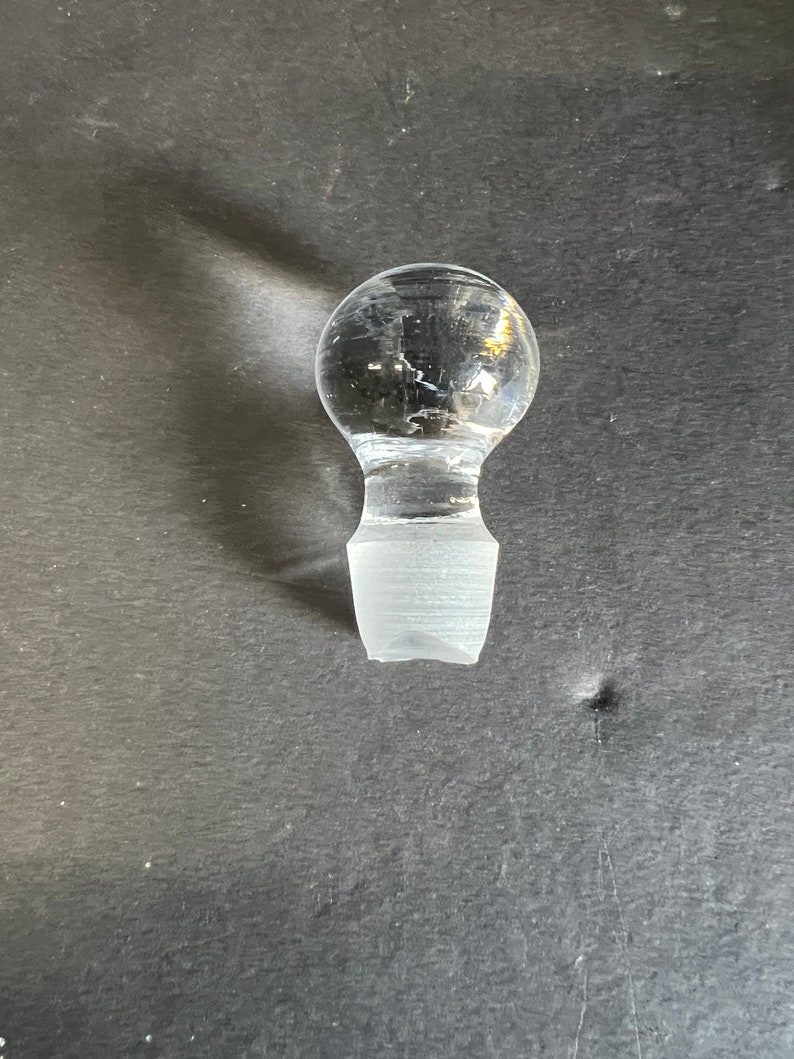May include: A clear glass bottle stopper with a round top and a flat, rectangular base.