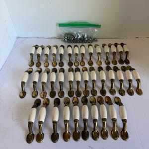 May include: A collection of thirty-three cabinet pulls featuring a white ceramic center handle and antique brass-toned metal ends. Each pull has a curved design with two mounting points. A plastic bag containing metal screws is included.