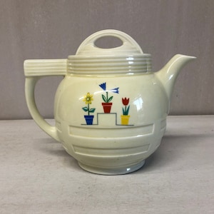May include: A white ceramic teapot with a floral design. The teapot has a round body with a lid and a handle. The floral design features three potted plants with flowers in yellow, red, and blue.