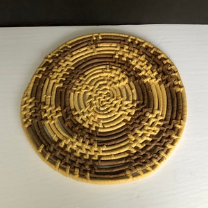 Puede incluir: Un posavasos redondo tejido con un patrón marrón y beige. El posavasos está hecho de materiales naturales y tiene un aspecto rústico.