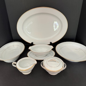 May include: A set of white porcelain dinnerware with gold trim. The set includes a large oval platter, two bowls, a gravy boat with a matching saucer, a creamer, and a covered sugar bowl.