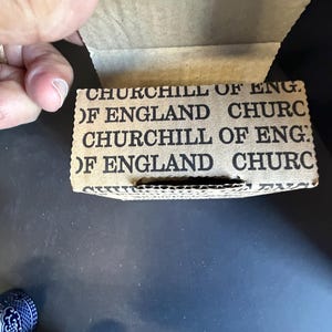 Peut inclure: Gros plan sur une bo&icirc;te en carton avec le texte "CHURCHILL OF ENGLAND" imprim&eacute; en noir. La bo&icirc;te est ouverte, r&eacute;v&eacute;lant l'int&eacute;rieur. La bo&icirc;te est tenue par une main. Le fond est une surface sombre.