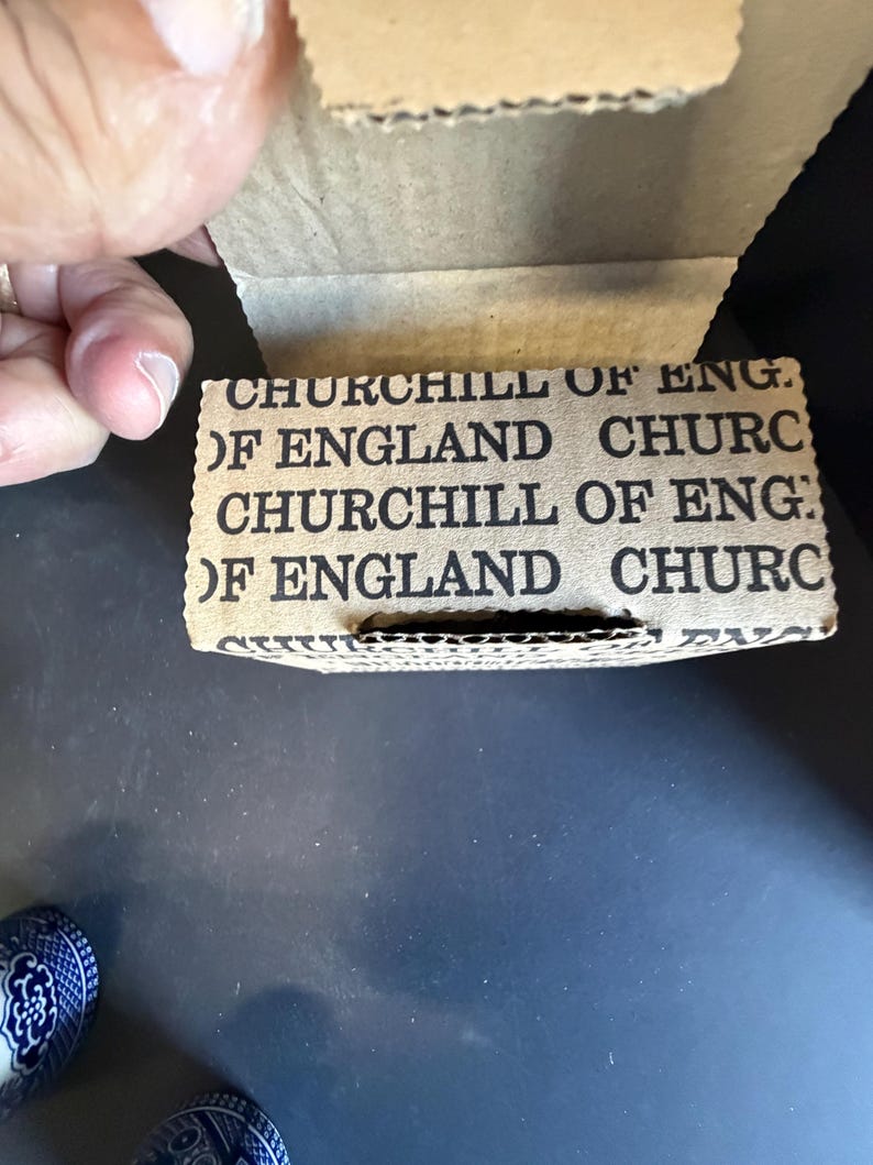 Peut inclure: Gros plan sur une bo&icirc;te en carton avec le texte "CHURCHILL OF ENGLAND" imprim&eacute; en noir. La bo&icirc;te est ouverte, r&eacute;v&eacute;lant l'int&eacute;rieur. La bo&icirc;te est tenue par une main. Le fond est une surface sombre.