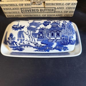 Peut inclure: Un beurrier en c&eacute;ramique bleu et blanc avec un couvercle orn&eacute; d'un motif Willow. Le plat repose sur un plateau assorti. La bo&icirc;te derri&egrave;re le plat indique "COVERED BUTTER" et "CHURCHILL OF ENGLAND".