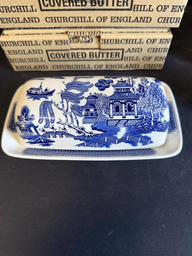 Peut inclure: Un beurrier en c&eacute;ramique bleu et blanc avec un couvercle orn&eacute; d'un motif Willow. Le plat repose sur un plateau assorti. La bo&icirc;te derri&egrave;re le plat indique "COVERED BUTTER" et "CHURCHILL OF ENGLAND".