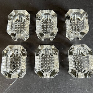 May include: Six clear glass salt cellars with a decorative, geometric design. The salt cellars are octagonal in shape and have a raised, textured center.