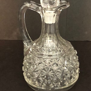 May include: A clear glass bottle with a textured surface and a round stopper. The bottle has a handle and a wide base.