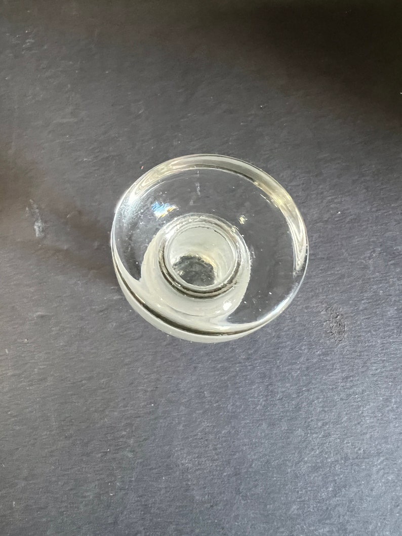 May include: A clear glass bottle stopper with a wide, flat top and a smaller, round opening in the center.