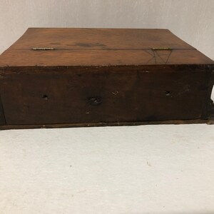 May include: A wooden box with a hinged lid and two brass hinges. The box is closed and sitting on a white surface.
