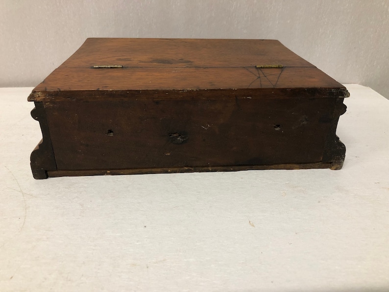 May include: A wooden box with a hinged lid and two brass hinges. The box is closed and sitting on a white surface.