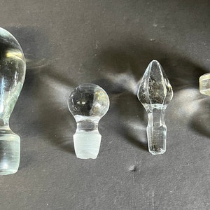 May include: Four glass bottle stoppers in different shapes and sizes. The stoppers are clear glass and have a variety of designs.