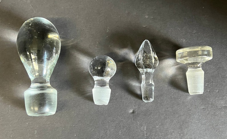 May include: Four glass bottle stoppers in different shapes and sizes. The stoppers are clear glass and have a variety of designs.