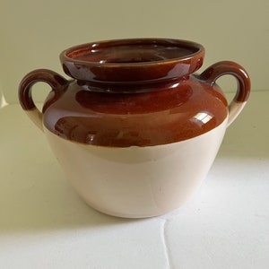 May include: A brown and white ceramic bean pot with two handles. The pot is glazed and has a smooth finish.