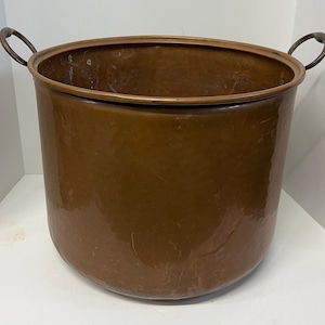 May include: A large, copper pot with two handles. The pot is round and has a shiny, slightly tarnished surface.