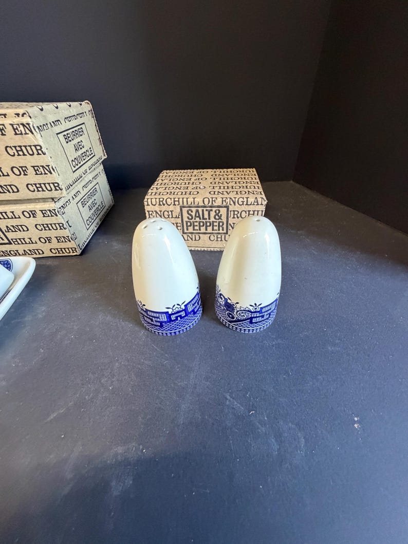 Peut inclure: Deux sali&egrave;res et poivri&egrave;res en c&eacute;ramique blanche avec des bandes &agrave; motifs bleus. Les sali&egrave;res et poivri&egrave;res sont &agrave; c&ocirc;t&eacute; d'une bo&icirc;te qui indique "Churchill of England Salt & Pepper". Les bo&icirc;tes sont blanches avec du texte noir.