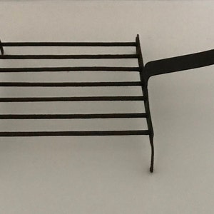 May include: Black metal fireplace grate with a handle. The grate has six horizontal bars and two vertical legs.