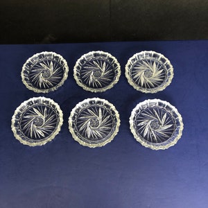May include: Six clear glass coasters with a cut-out starburst design. The coasters are arranged in two rows of three on a blue surface.