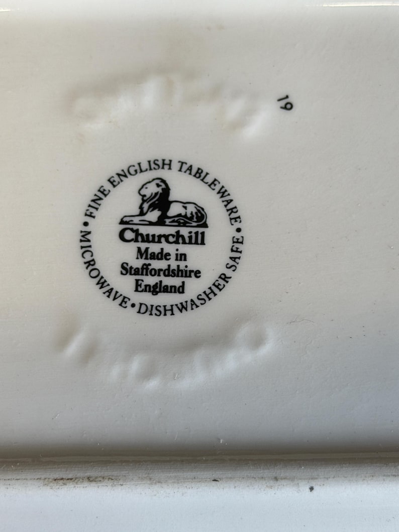 Peut inclure: Gros plan sur une assiette en c&eacute;ramique blanche avec le texte "FINE ENGLISH TABLEWARE" et "Churchill Made in Staffordshire England" dans un motif circulaire noir. L'assiette est adapt&eacute;e au micro-ondes et au lave-vaisselle.