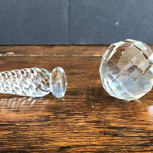 May include: Two clear glass bottle stoppers. One stopper is a long, thin, twisted design. The other stopper is a short, thick, faceted design.