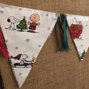 May include: A festive Christmas banner featuring Snoopy and Charlie Brown characters. The white pennants are decorated with holiday scenes and snowflakes, trimmed with red and green ribbons. The banner is strung across a neutral background.