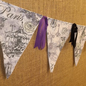 May include: A fabric bunting banner featuring triangular flags with a light gray and white print of Paris landmarks, roses, and script text. The banner is accented with alternating black and purple ribbon ties attached between the fabric flags.