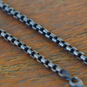 May include: A silver chain necklace with a box chain design. The chain is on a wooden surface.