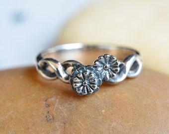 Könnte beinhalten: Ein silberner Ring mit einem verdrehten Band und zwei Blumendesigns.