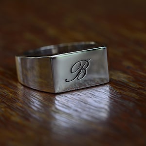 Peut inclure: Une bague chevalière en métal argenté poli avec une face rectangulaire plate gravée de la lettre B en écriture cursive. La bague présente une surface métallique lisse et réfléchissante ainsi qu'un anneau large qui s'affine vers l'arrière.