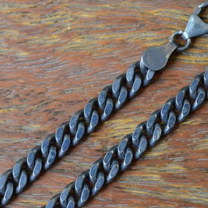 Puede incluir: Un collar de cadena de eslabones tipo barbada en plata oxidada de color gris oscuro, con eslabones ovalados entrelazados, bordes pulidos y un cierre de mosquetón. La cadena metálica presenta un aspecto mate y texturizado con oxidación.