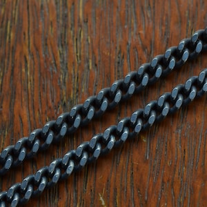 May include: A black metal chain with a curb link pattern. The chain is lying on a wooden surface.