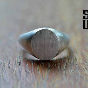 May include: A silver-toned signet ring with a smooth, circular, brushed-finish face. The ring features a solid band that tapers toward the back. The text SOUL is printed in black in the upper right corner of the image.