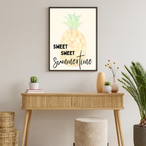 Può includere: Una stampa incorniciata con un'illustrazione ad acquerello di un ananas e il testo "Sweet Sweet Summertime". La stampa è appesa a una parete bianca sopra un tavolo consolle in legno.